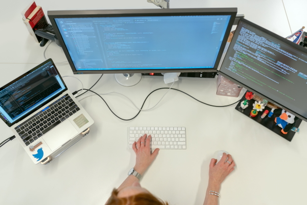 de handen van een vrouw aan een bureau met voor zich drie schermen en een toetsenbord